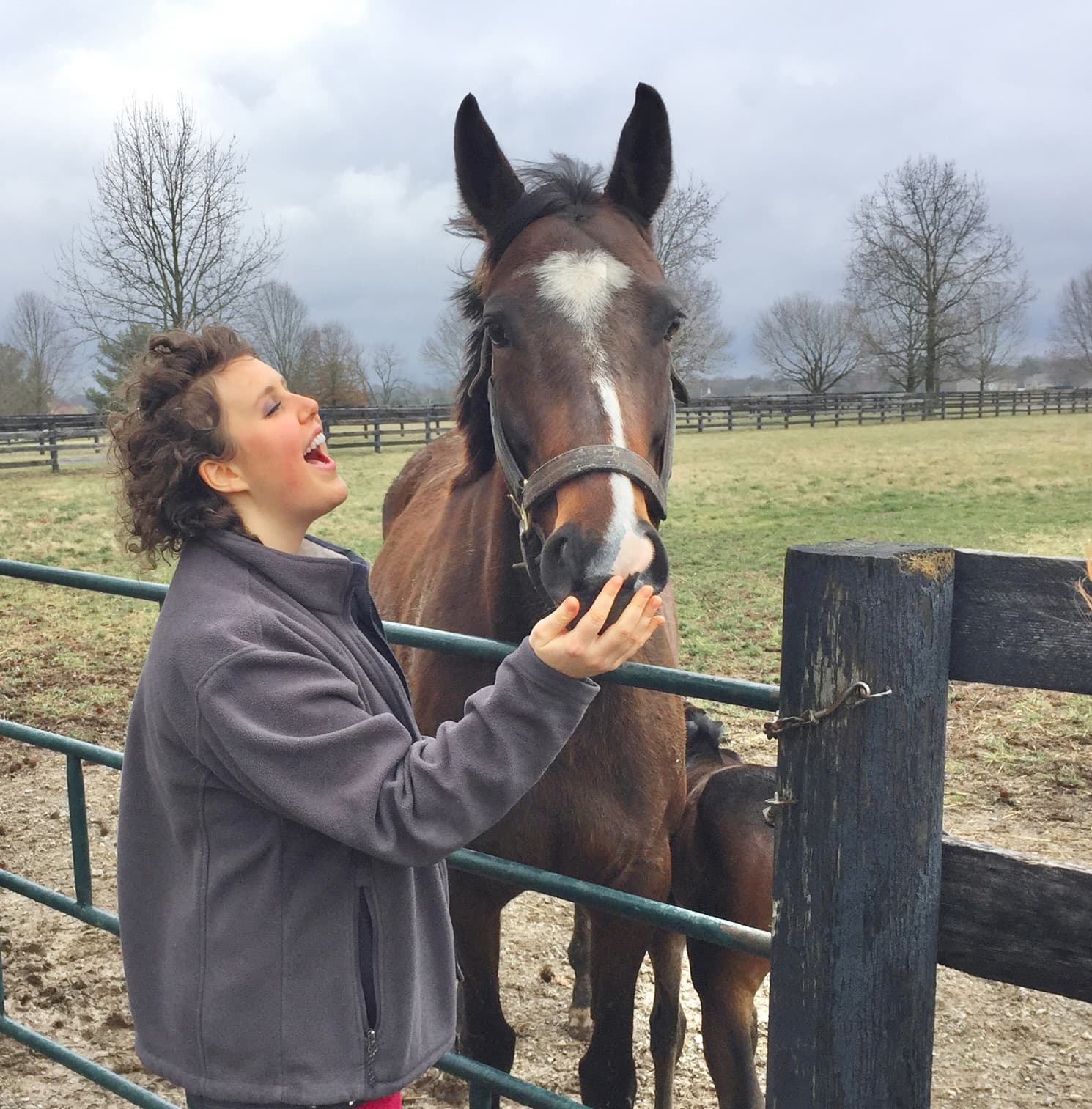 A woman smiling and playfully interacting with a brown horse at a farm.