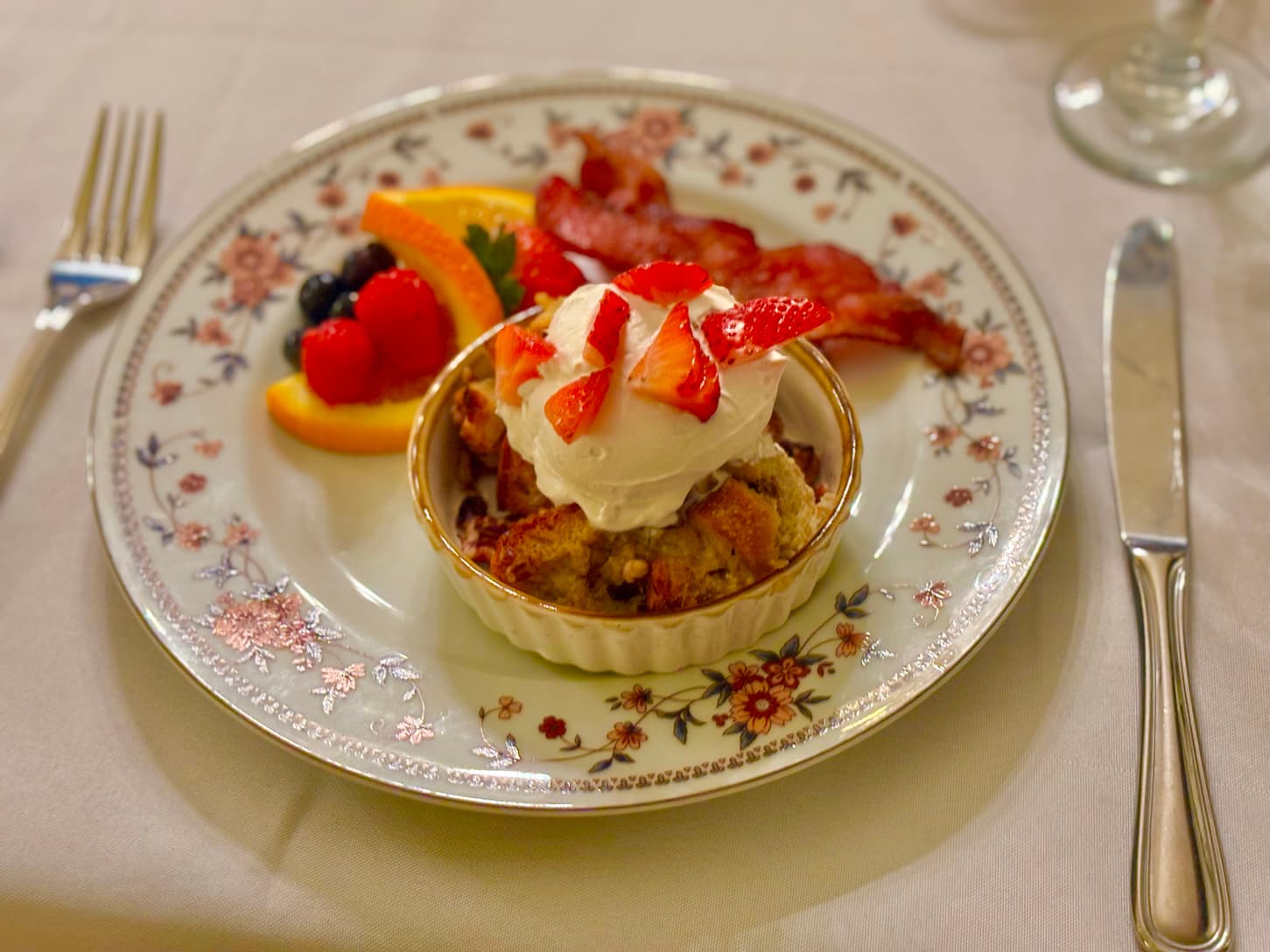 A dessert with whipped cream and strawberries, served with a side of fruit and bacon on a decorative plate.