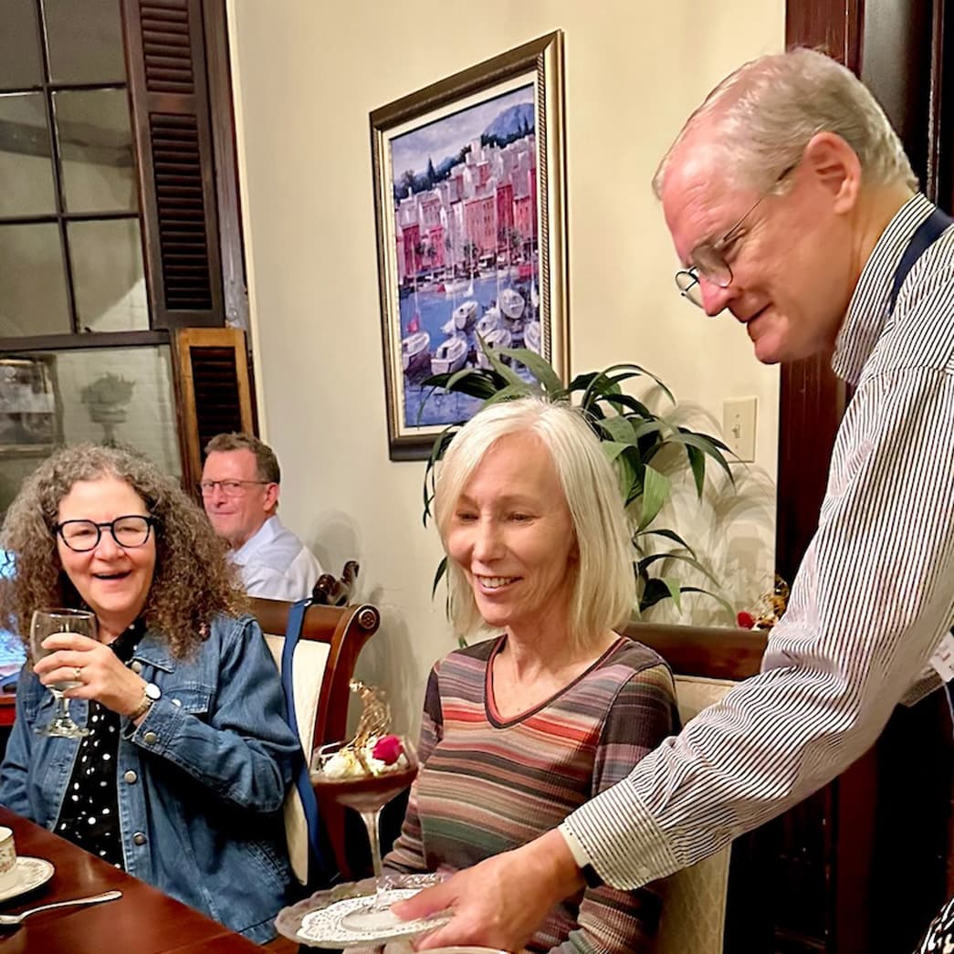A group of four people enjoys a meal together, with one person serving food at a dining table.